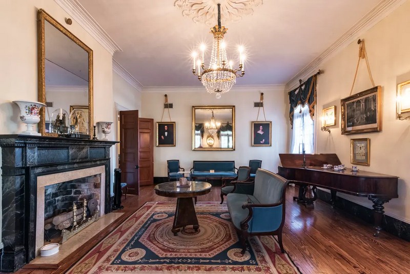 Дверь с панелями из кипарисового дерева открывается из прихожей особняка в гостиную. С большого декоративного медальона на потолке гостиной свисает изысканный акцент: хрустально-латунная люстра с круглым канделябром. Пол покрыт полированной сосной, а каминная полка окрашена под черно-белый мрамор. Синяя бархатная мебель во французском стиле Карла X и Луи-Филиппа обеспечивают места для отдыха в этой комнате. (Oak Alley Foundation)