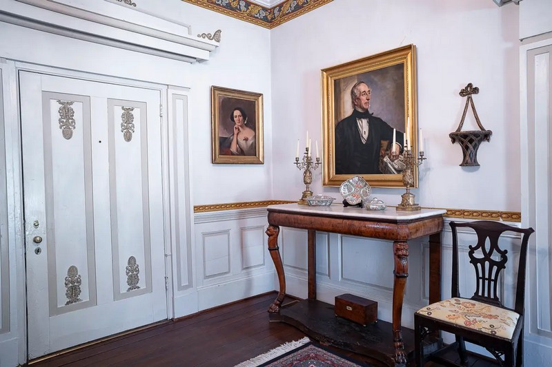 В прихожей дома висит копия портрета Джона Тайлера в 1859 году, написанного художником Джорджем Питером Александром Хили (1813–1894); оригинал картины находится в Национальной портретной галерее в Вашингтоне. На меньшей картине изображена родственница Джона Тайлера: Джейн Кэри Гаррисон Руффин. Широкая, окрашенная входная дверь украшена аппликацией в стиле греческого возрождения, вдохновенной книгой по дизайну 19 века архитектора Минарда Лефевера (1798–1854). «Это прикосновение Джулии Гардинер Тайлер, который она добавила в дом», — сказала исполнительный директор Анник Даннинг. Вдоль панельных перил стульев – кайма из обоев с шестиугольным узором. (Sherwood Forest Plantation Foundation)