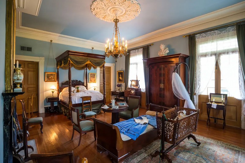 Каждая из главных комнат на плантации Oak Alley имеет исключительный и неповторимый медальон на потолке, с которого свисает не менее уникальный светильник. Спальня на верхнем этаже не исключение. Густо обставленная антикварной мебелью 19 века, изготовленной вручную, центральными элементами этой комнаты являются кровать-диван в форме саней и изящно резная люлька на точеной основе с ножками в стиле Чиппендейл. Резная кровать с балдахином обита лесно-зеленым бархатом с золотыми кистями, свисающими с верхней рамы кровати. (f11photo/Shutterstock)