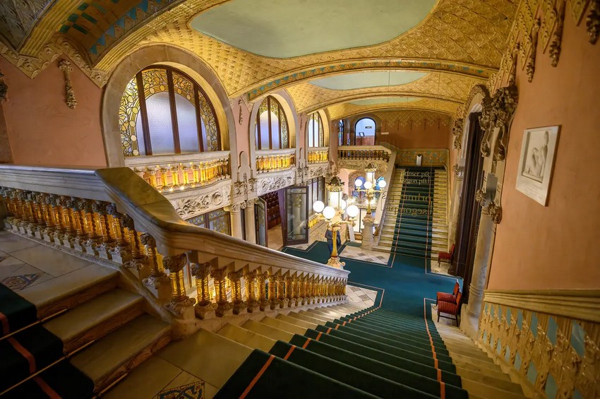 Великі мармурові сходи, обрамлені коронованими лампами на колонах, мають мармурову балюстраду, що підтримується прозорими жовтими скляними балясинами. Фойє та сходи є прикладом елементів каталонського модерну з плавними та вигнутими лініями, квітковими мотивами, вітражами та органічними зеленими та золотими кольорами. (Mitzo/Shutterstock)