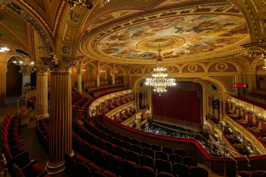 Триповерховий зал у формі підкови пропонує відвідувачам справжнє театральне видовище червоних і золотих відтінків, вражаючу фреску на стелі та бронзово-скляну люстру, що висить у центрі купола. Спочатку люстра освітлювалася газом, але зараз ця майже тритонна конструкція освітлюється електрикою за допомогою понад 200 лампочок. Розкішні позолочені коринфські колони по всій аудиторії підтримують прикрашені арки. (posztos/Shutterstock)