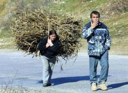 Чоловік іде, покурюючи цигарку, а позаду нього жінка несе оберемок сіна