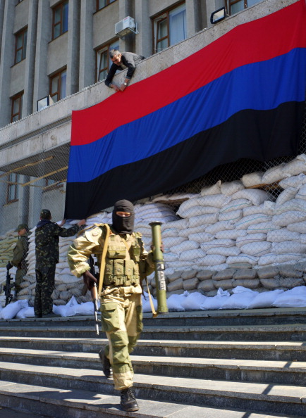 Озброєний чоловік у військовій формі проходить повз проросійських активістів, які вивішують прапор так званої «Народної Республіки Донбасу» на адмінбудівлі в м. Слов'янськ, 21 квітня 2014 р. Фото: GENYA SAVILOV/AFP/Getty Images