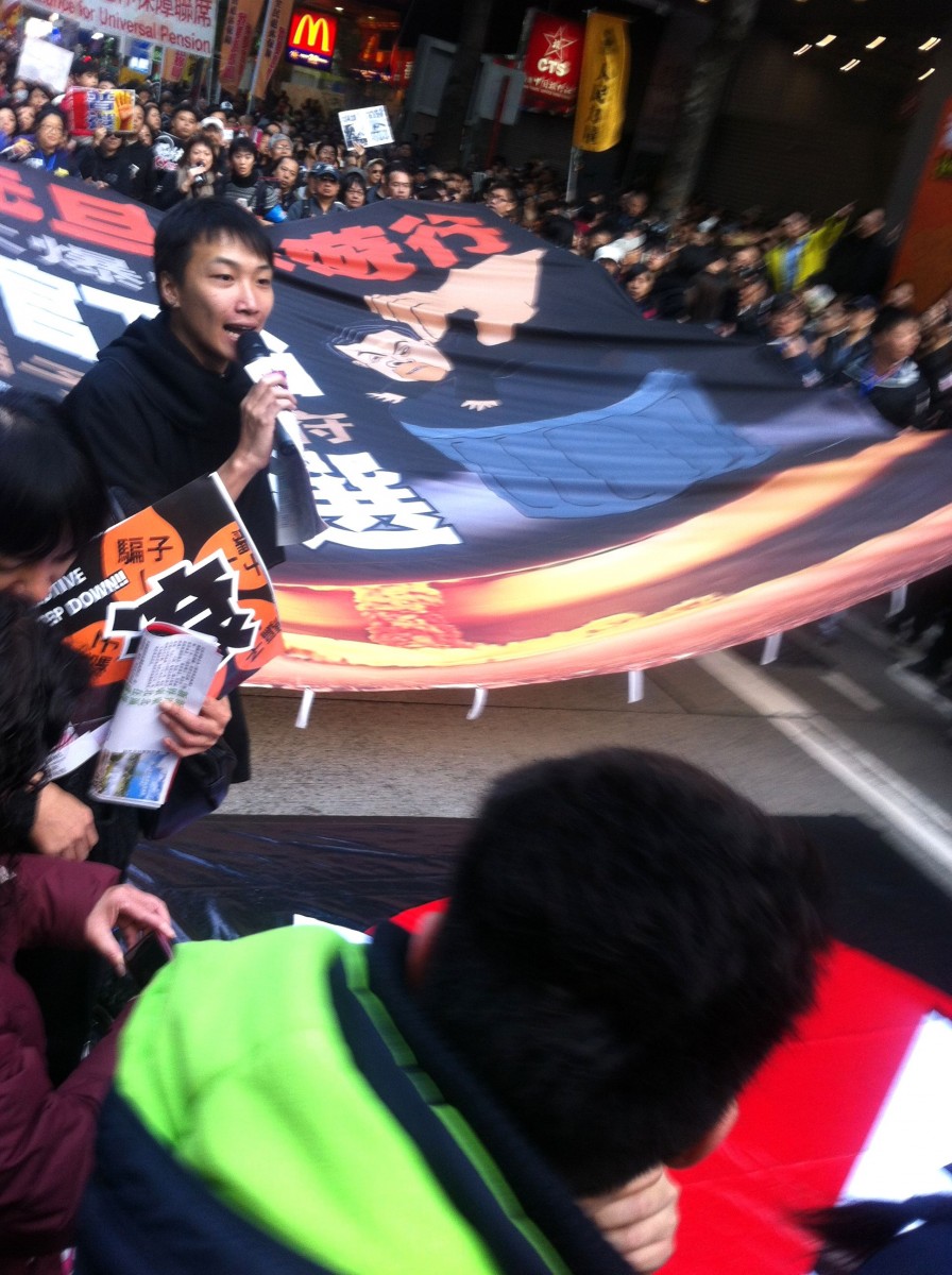 Группа жителей города держит баннер с надписью: «Новогодний парад». На баннере в перечеркнутом красном круге изображены коммунистические символы, а также большая рука, выбрасывающая манекен Лян Чженьина в мусорную корзину