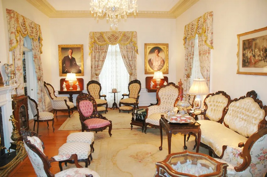 В гостиной стоит резная мебель в викторианском стиле, обитая гобеленом с цветочным принтом. Люстра свисает с потолка, окаймленного золотым лепным покрытием, которое оттеняет золотой антикварный ковер, цветочные парчовые драпировки в форме водопада и латунную каминную полку. Золотые рамы оттеняют картины Беллингратов Джея Джейкобса, того самого художника, нарисовавшего официальный портрет президента Гарри Трумэна в Белом доме. (Bellingrath Gardens & Home)